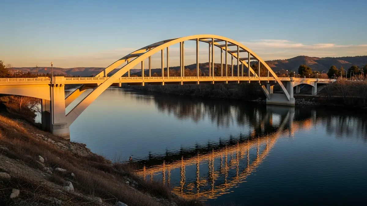 A beautiful professional landscape photograph of the Johnny Cash Overpass in Folsom, California, spanning the American River at golden hour. The bridge's elegant cream structure glows under warm, sun-drenched light, reflected in the deep blue river below. Distant hills show subtle rust and terracotta hues.