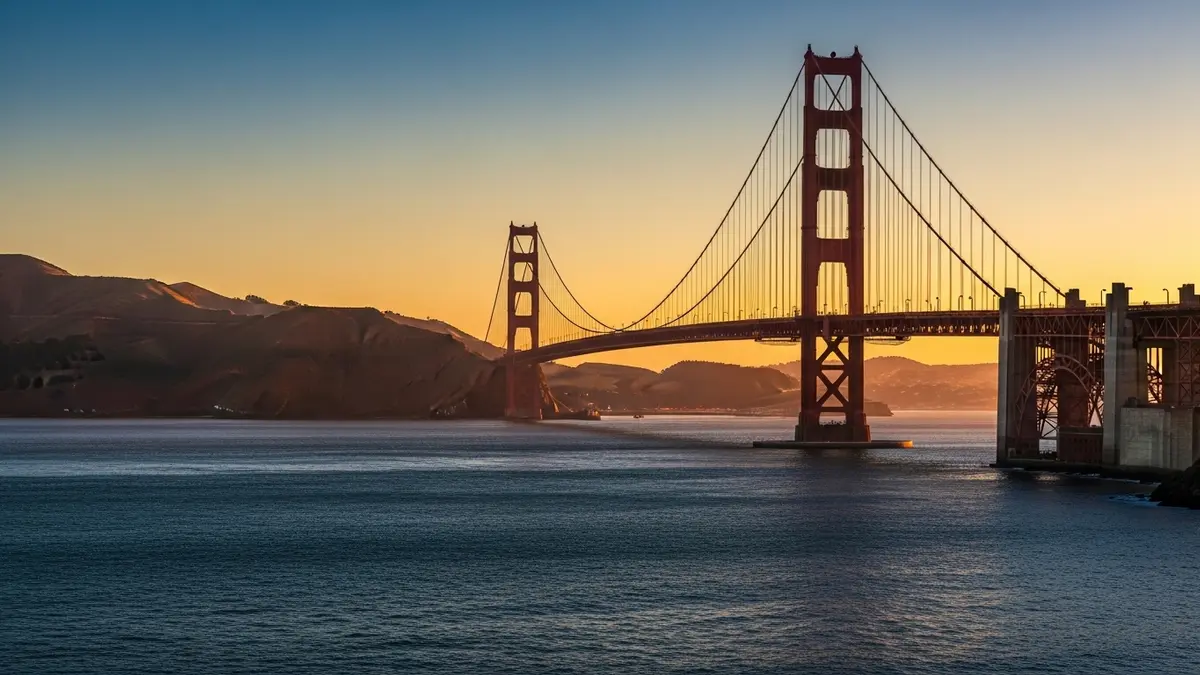 A professional, high-quality landscape photograph of the Golden Gate Bridge in San Francisco, CA, bathed in warm golden hour light. The iconic terracotta-red bridge stands prominently over the deep navy-blue waters of the bay, with the distant city skyline and rolling hills gently illuminated by the setting sun's creamy, amber glow.