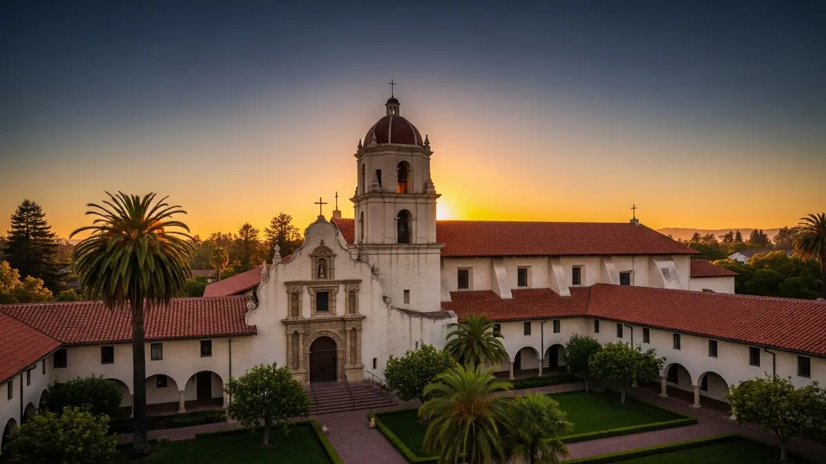 A beautiful, sun-drenched view of Mission Santa Clara de Asís on the Santa Clara University campus in Santa Clara, CA, during golden hour. The historic architecture features warm terracotta roof tiles and creamy stucco walls, framed by lush green landscaping under a deep blue and orange sky, evoking a serene and elegant atmosphere.