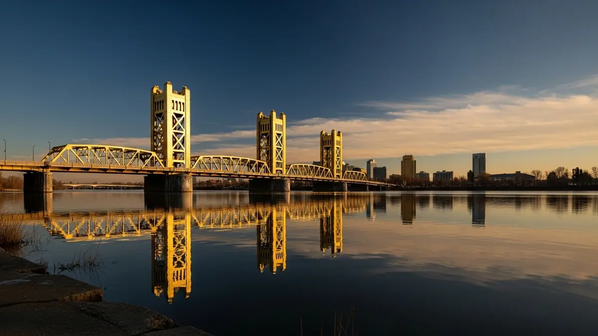 The iconic golden Tower Bridge viewed from West Sacramento at sunset. The calm Sacramento River reflects the warm, golden light and the deep blue sky, with the Sacramento skyline visible in the distance.